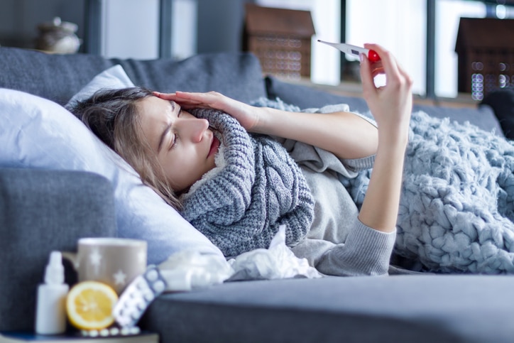 Young woman with fever and headache is measuring temperature with thermometer, treated at home.